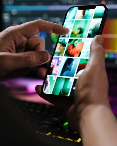 Close up of a white man hand holding mobile phone over computer screen on a table indoors