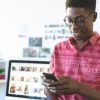 Front view of happy young African-American graphic designer using mobile phone at desk in office