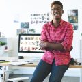 Portrait of young African-American graphic designer looking and smiling at camera while sitting at desk in office